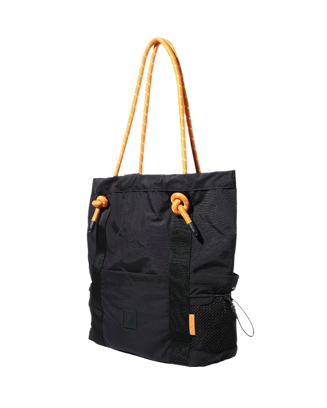 Three-quarter angled view of the black SYMA Noble minimalist everyday tote bag, showing the front pocket, side mesh water bottle pocket, 2.5cm vertical webbing, and orange paracord straps