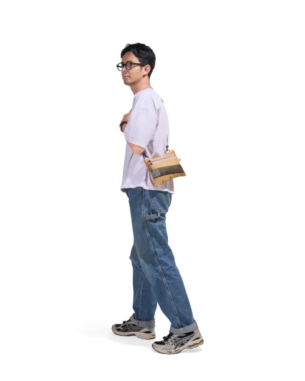 Full-body lifestyle profile of a male model in casual streetwear wearing the SYMA Mantra khaki sacoche bag across his body, demonstrating its ultra-slim 1cm profile and compact everyday carry utility
