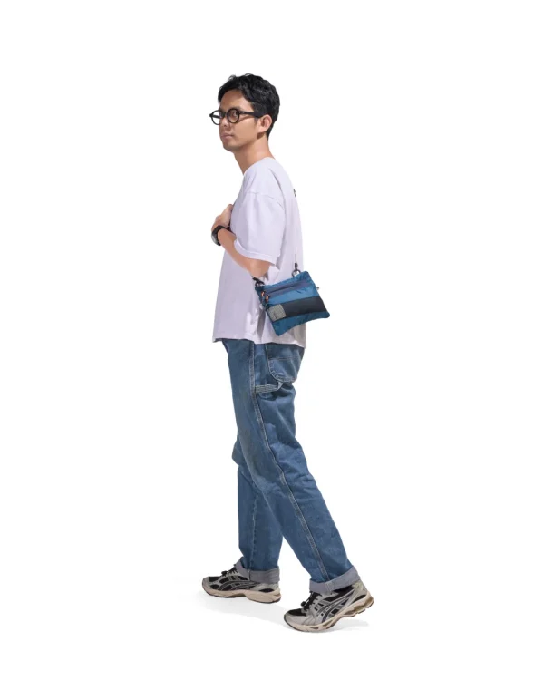 Full-body lifestyle profile of a male model in casual streetwear wearing the SYMA Mantra navy sacoche bag across his body, demonstrating its ultra-slim 1cm profile and compact everyday carry utility