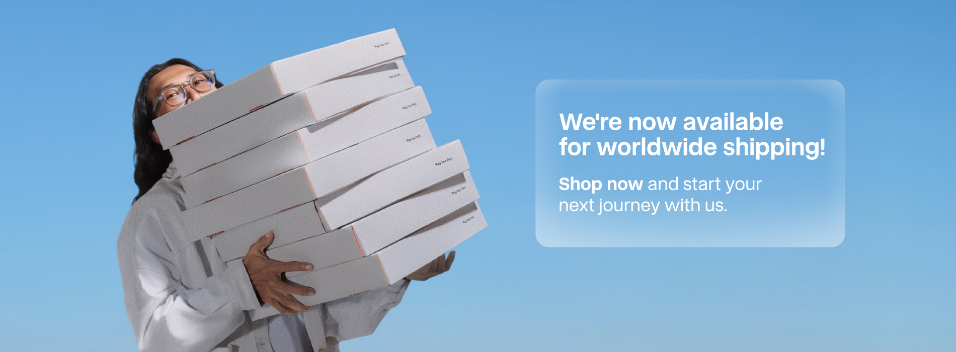 Banner announcing SYMA worldwide shipping, showing a person carrying a large stack of white shipping boxes against a clear blue sky, with text reading: We're now available for worldwide shipping! Shop now and start your next journey with us.