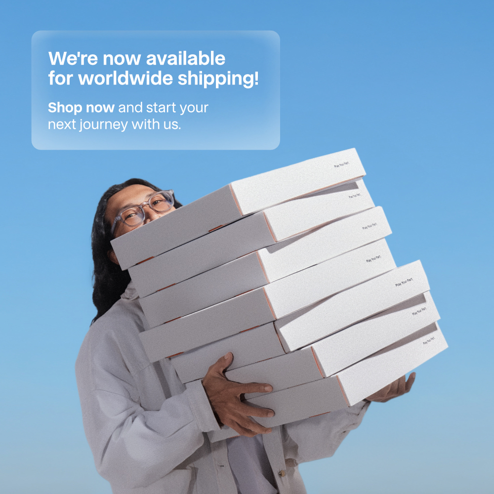 Mobile banner announcing SYMA worldwide shipping, showing a person carrying a large stack of white shipping boxes against a clear blue sky, with text reading: We're now available for worldwide shipping! Shop now and start your next journey with us.